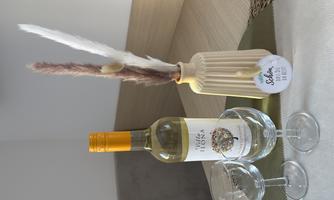 A bottle of white wine stands next to two glasses on a table. There is also a decorative vase with dried flowers.