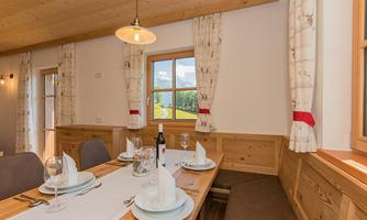 A bright dining area with a wooden table, decorated with dishes and glasses. Through the windows, you look out onto a green landscape.