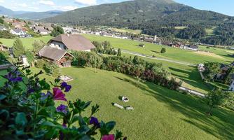 A wide landscape with green meadows and mountains in the background. In the foreground, flowers and a spacious garden can be seen.