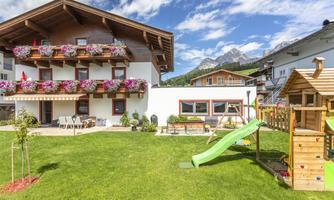A cozy house with flowers in the windows and a well-kept garden. In the background, there are mountains and a playground with a slide.