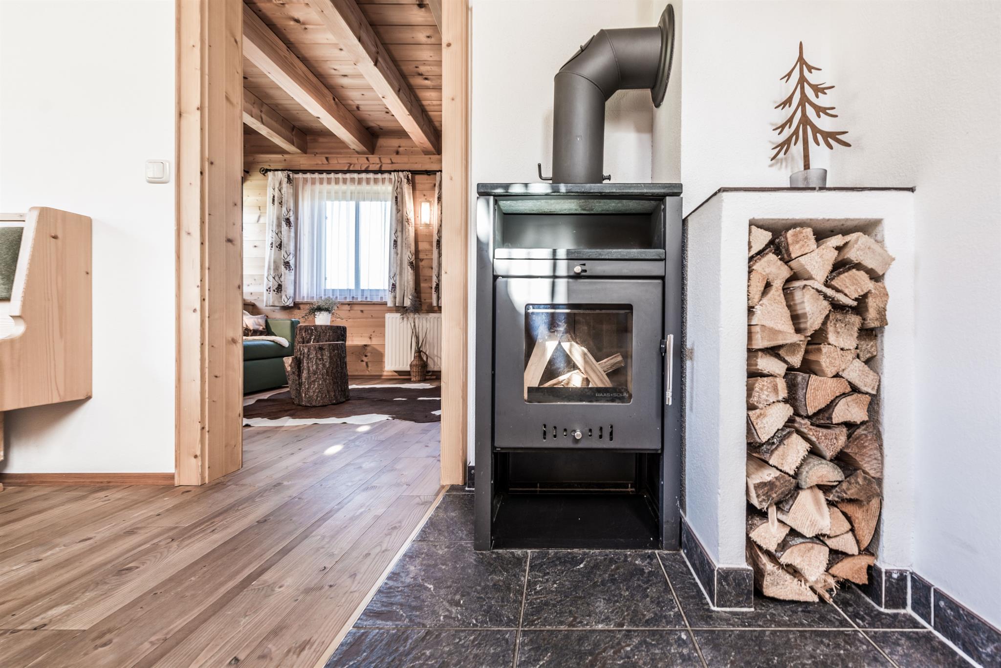 A cozy room with a wood stove and a stack of wood nearby. The floor is made of wood and the room exudes a warm atmosphere.