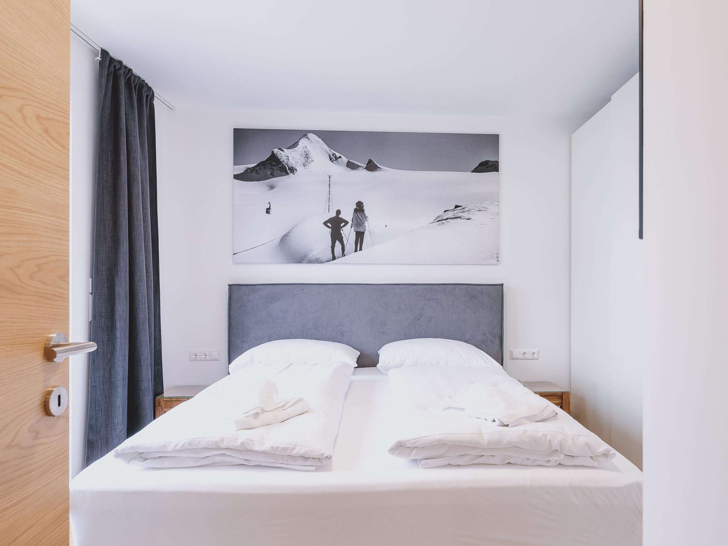 A modern bedroom with a double bed and a large wall mural of snow-covered mountains. The room is bright and minimally furnished.