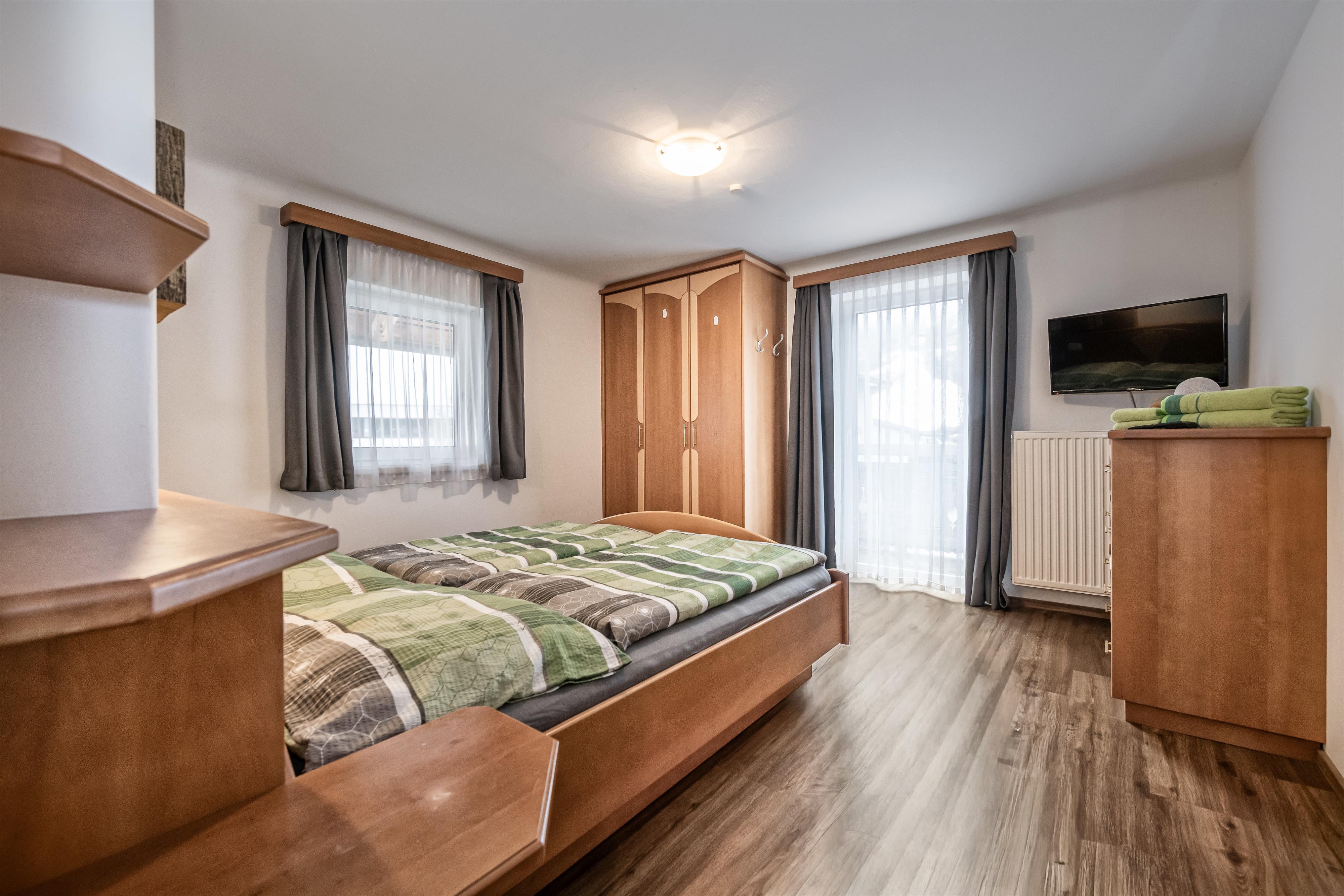 A cozy bedroom with a double bed and wooden shelves. The room has large windows and plenty of natural light.