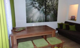 A modern living room with a wooden table and green seat cushions. A large picture of a forest with rays of light hangs on the wall.