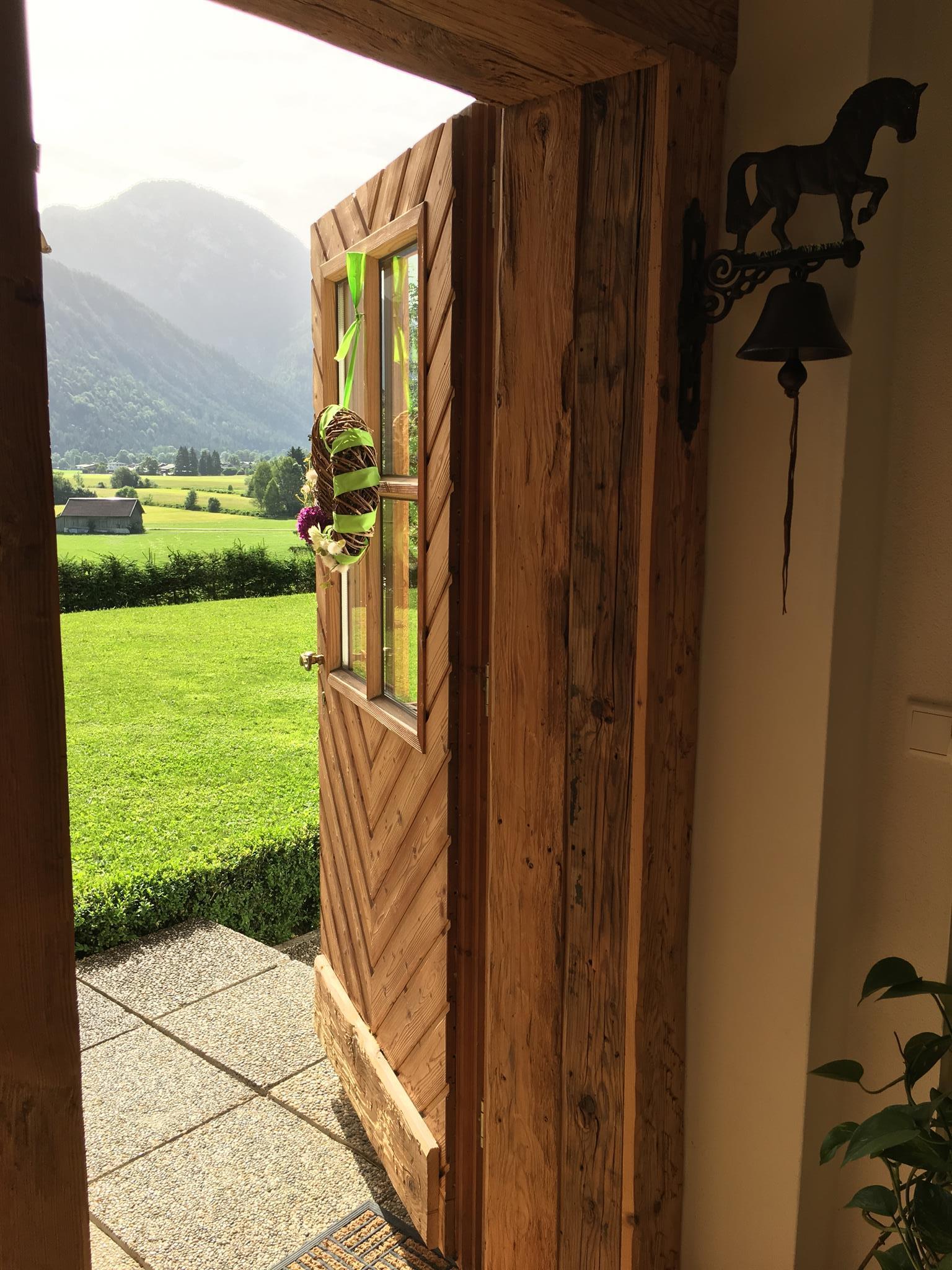 A wooden door stands open, offering a view of a green meadow and hills in the background. A wreath hangs on the door, and a hoof bell holder is visible.