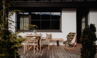A cozy outdoor area with a wooden table and chairs. In the background, there are large windows and green plants visible.