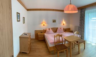 A cozy bedroom with two beds and warm wood paneling. There is a table, chairs, and a large window with plenty of natural light.