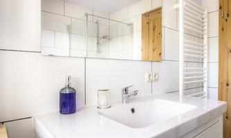 A modern bathroom with a white vanity top and a large mirror. Decorative objects such as a blue bottle and a glass are on the shelf.