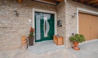 A modern front door in green with an elegant design. The entrance area is decorated with stone masonry and plants.