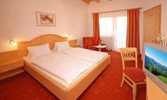 A cozy bedroom with a double bed, a desk, and an armchair. Large windows with curtains let in plenty of light.