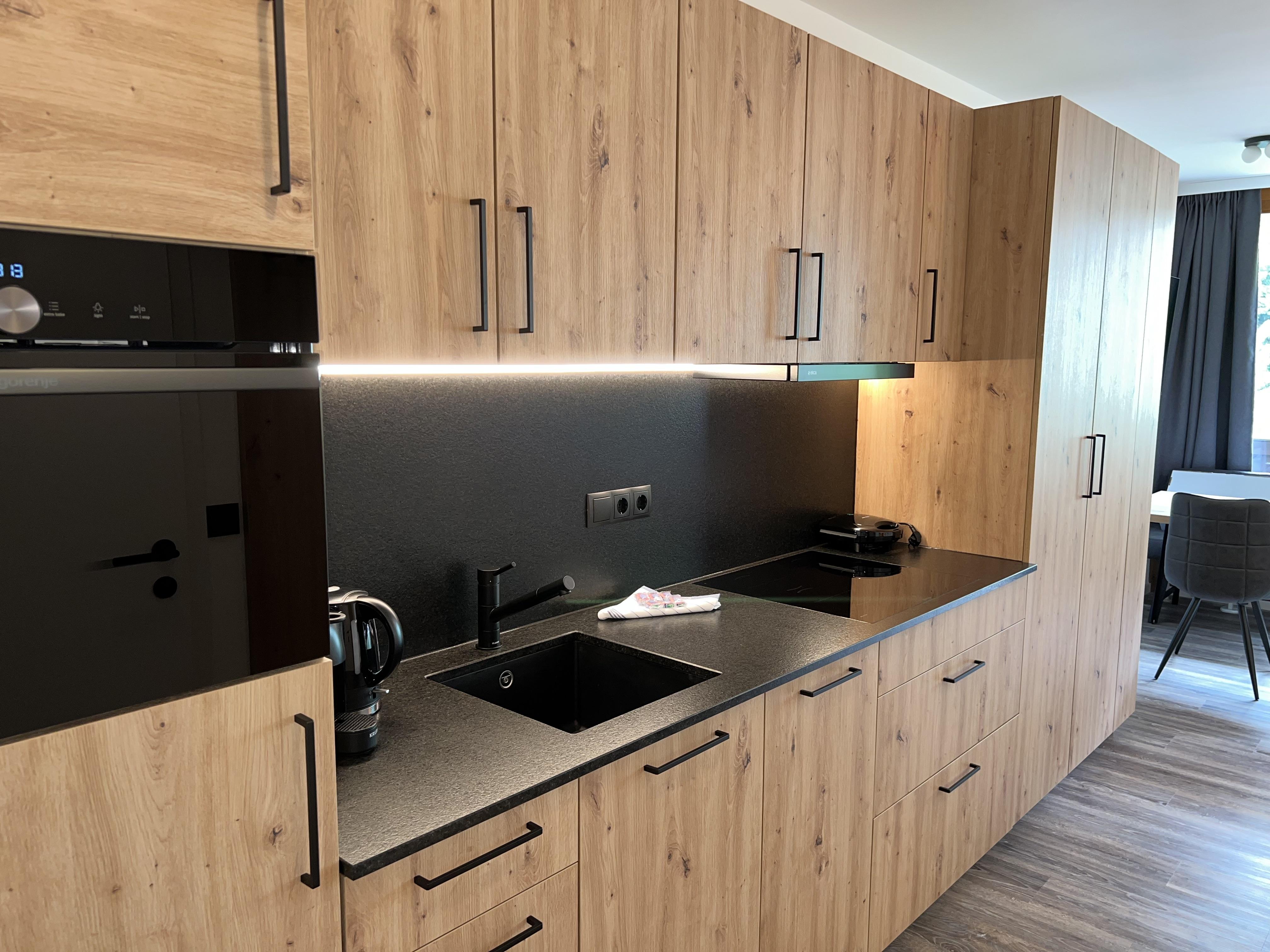 A modern kitchen with wooden cabinets and a black countertop. It is well equipped with a stove, kettle, and sink.