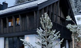 A modern wooden house in the snow, surrounded by snow-covered fir trees. The sky is gray and the atmosphere is calm and wintry.