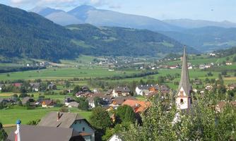 A picturesque village scene with beautiful houses and a church in the foreground. The gentle hills and mountains in the background create an idyllic landscape.