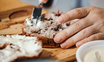 A hand is cutting a slice of bread and spreading it with a spread. In the foreground, there is a bowl with more cream visible.