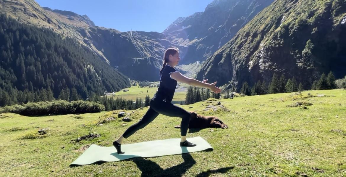 A woman is doing yoga on a mat in nature. In the background, green mountains and a clear sky can be seen.