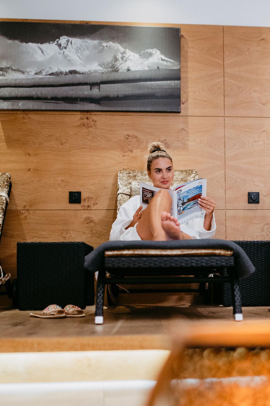 A woman in a bathrobe relaxes on a lounge chair and reads a magazine. In the background, wooden panels and a piece of art can be seen.