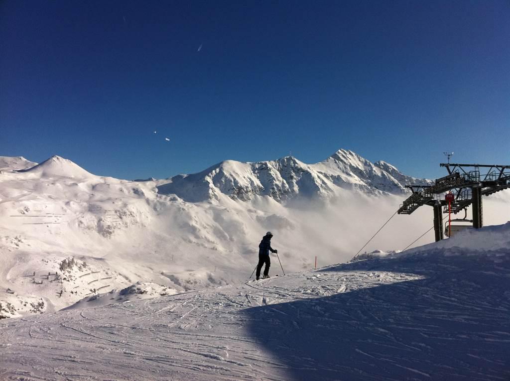 A snow-covered mountain landscape with a clear blue sky. A skier is going down the slope.