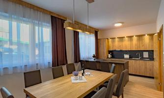 A modern kitchen with a large wooden dining table and six chairs. The room design is bright and friendly with large windows and warm wood tones.