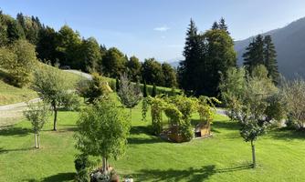 A green meadow with various trees and a cozy gazebo. In the background, gentle hills and a bright blue sky are visible.