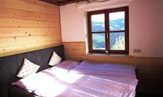 A cozy bedroom with wooden paneling and a large window. Bright blankets and pillows are on the bed.