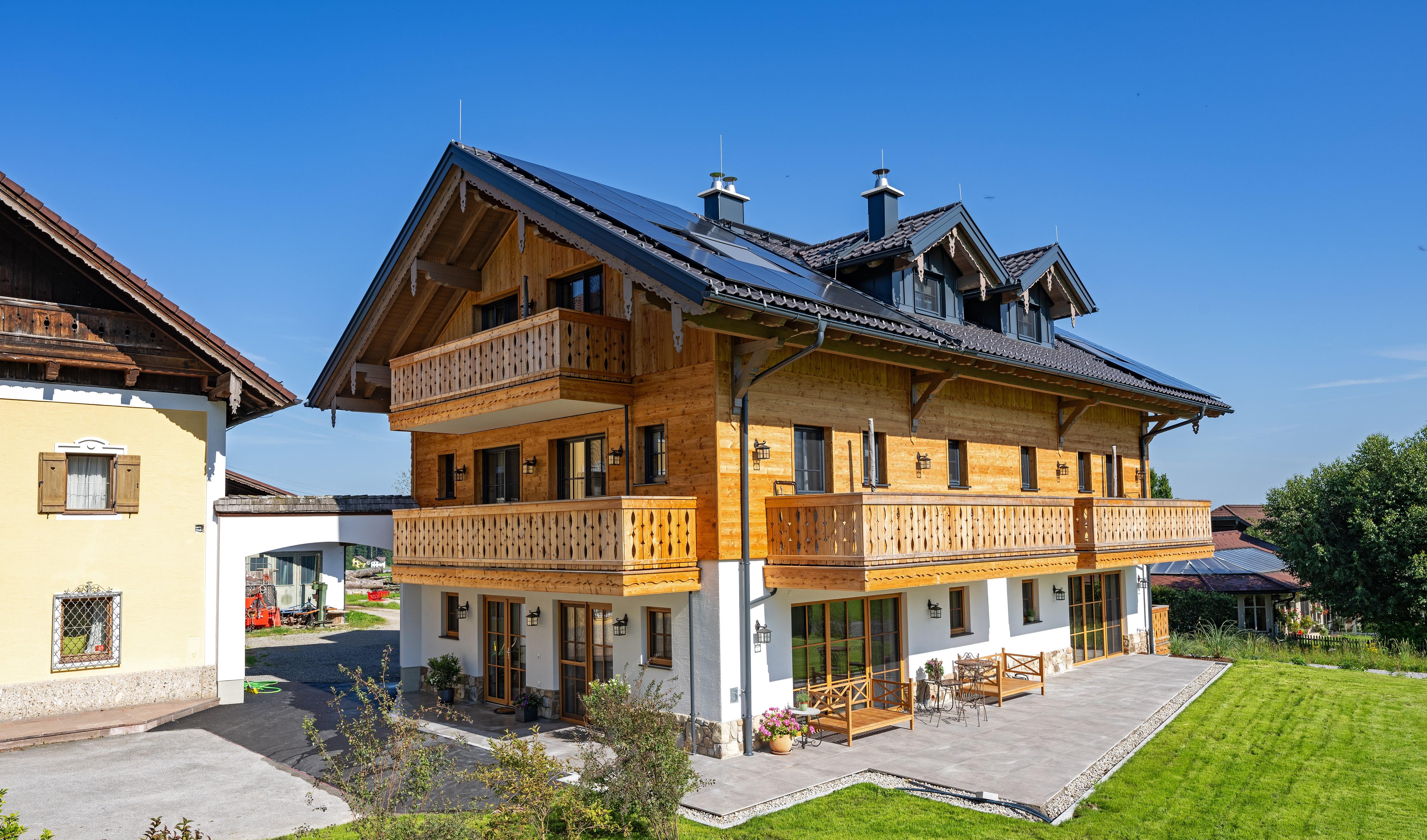 A modern wooden house with several balconies and large windows. The garden is well-maintained and surrounded by green scenery.
