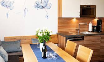 A modern dining room with a wooden table and a cozy seating area. On the table is a vase with white roses and a bottle of wine.