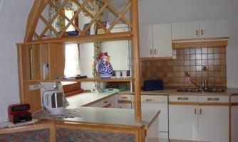 A bright kitchen with modern furniture and wooden shelves. The equipment includes a kettle, a cutlery corner, and an open passage bar.