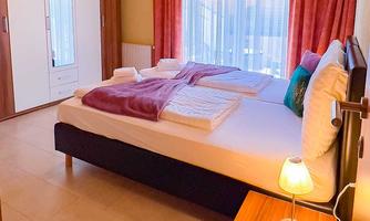 A bright bedroom with a large bed and soft pillows. Light curtains hang in front of the windows, and there is a wardrobe as well as a lamp on the nightstand.
