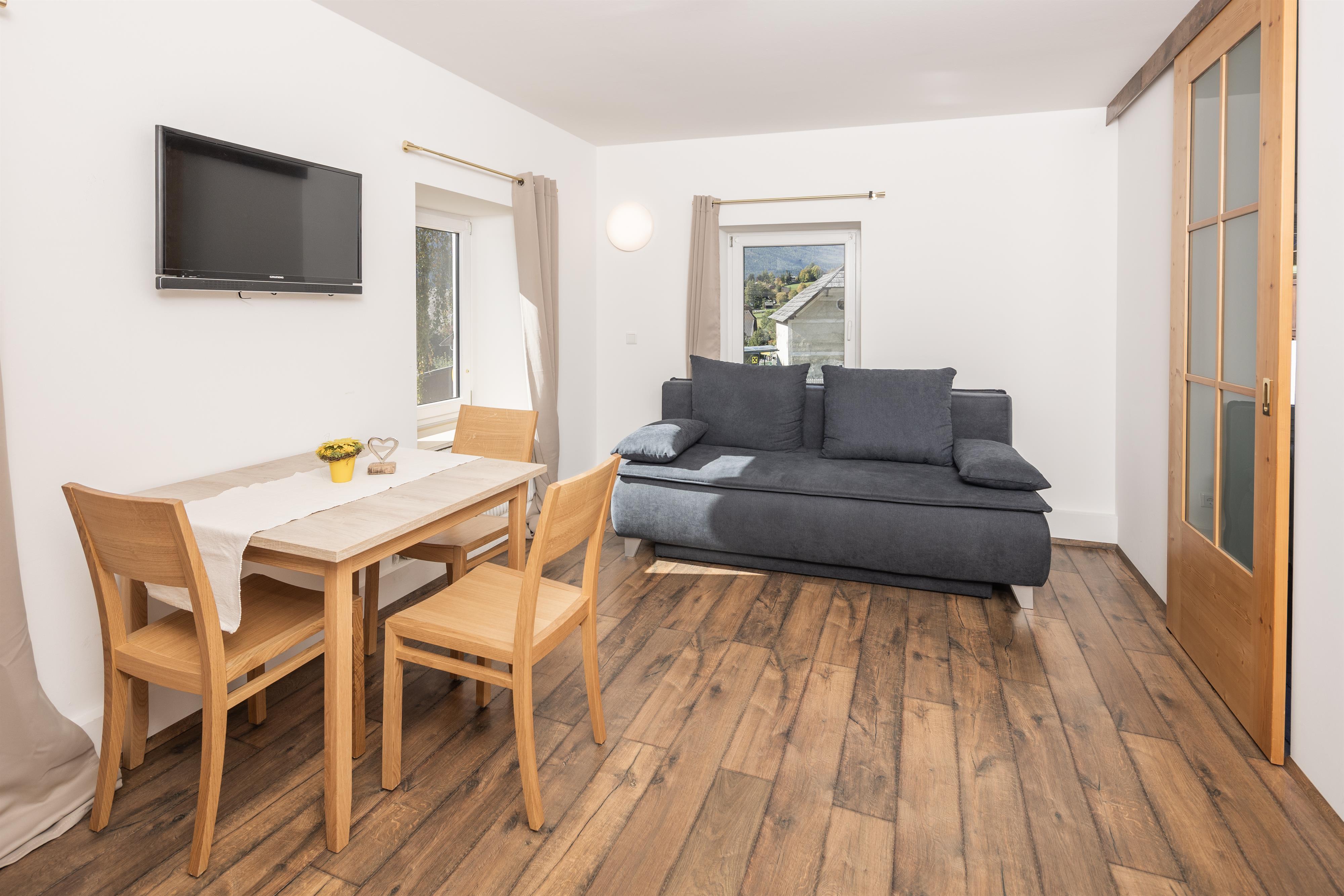 A bright living area with a gray sofa and a wooden dining table. A television is mounted on the wall and there are large windows that let in plenty of light.