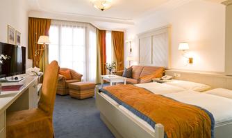 A modern hotel room with two beds and a cozy seating area. The decor is bright and inviting, with warm colors and ample daylight.
