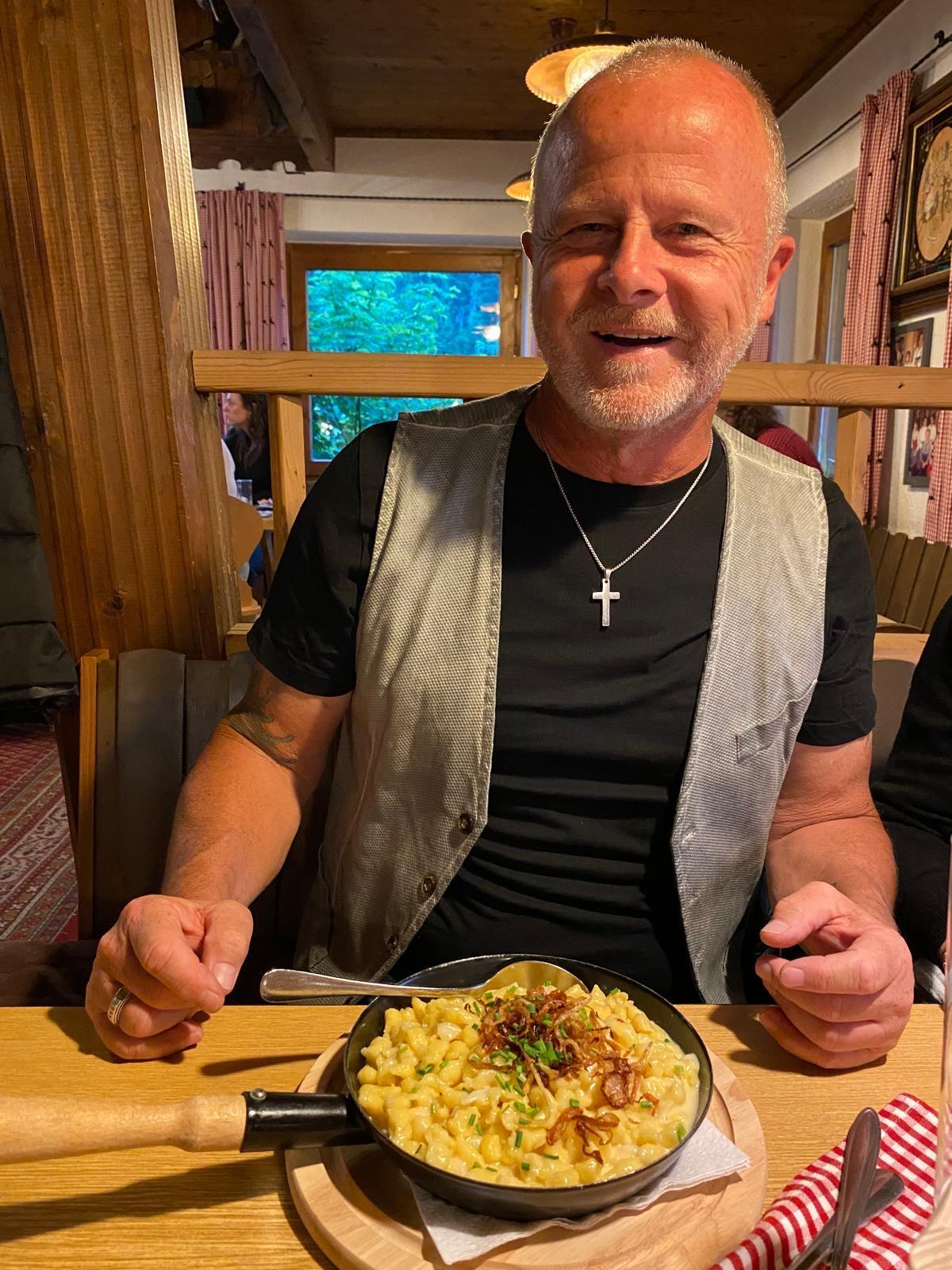 A smiling man is sitting at a wooden table, holding a pan with a dish in his hand. In the background, cozy restaurant elements and windows with a view of nature can be seen.
