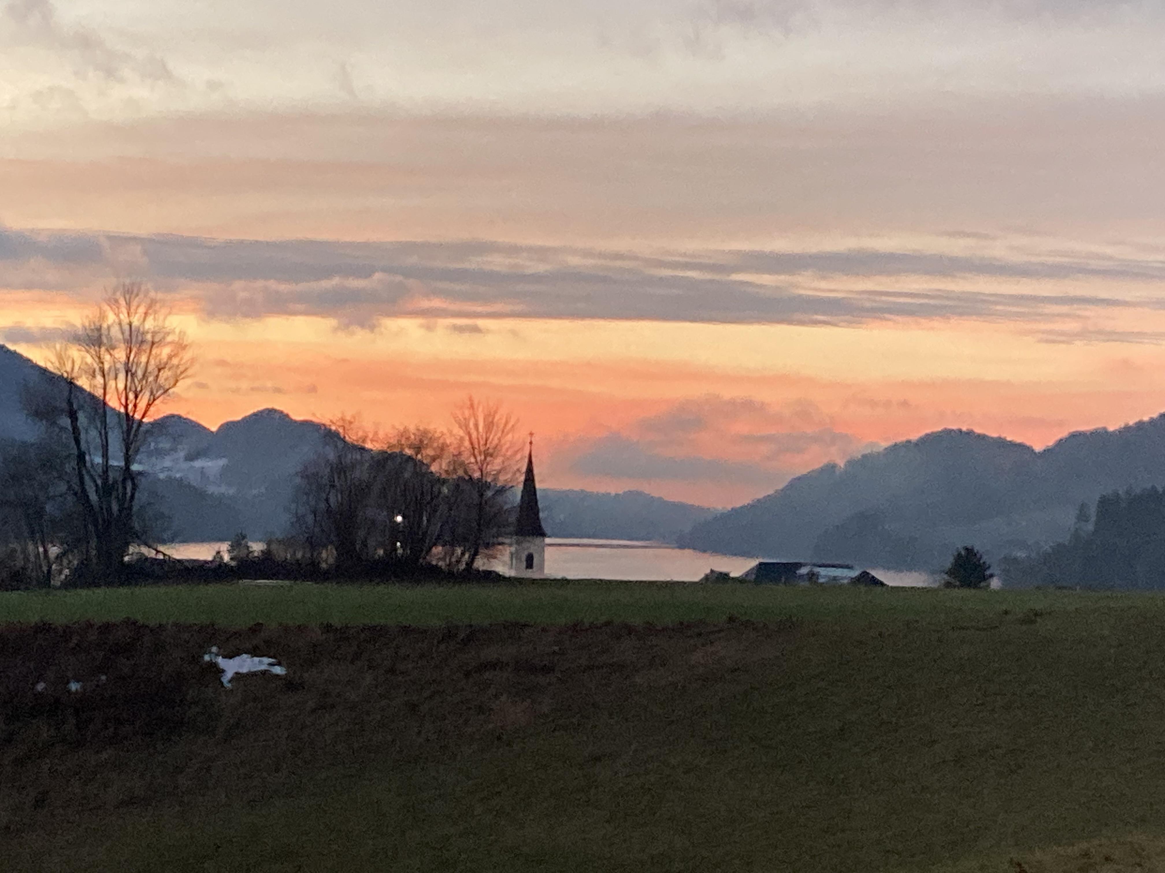 A peaceful sunset over a lake, surrounded by gentle hills. In the foreground, a small church with a pointed tower is visible.