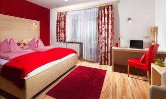 A modern bedroom with a red bed and warm wooden furniture. The room is bright and cozy, with a desk and a large window.