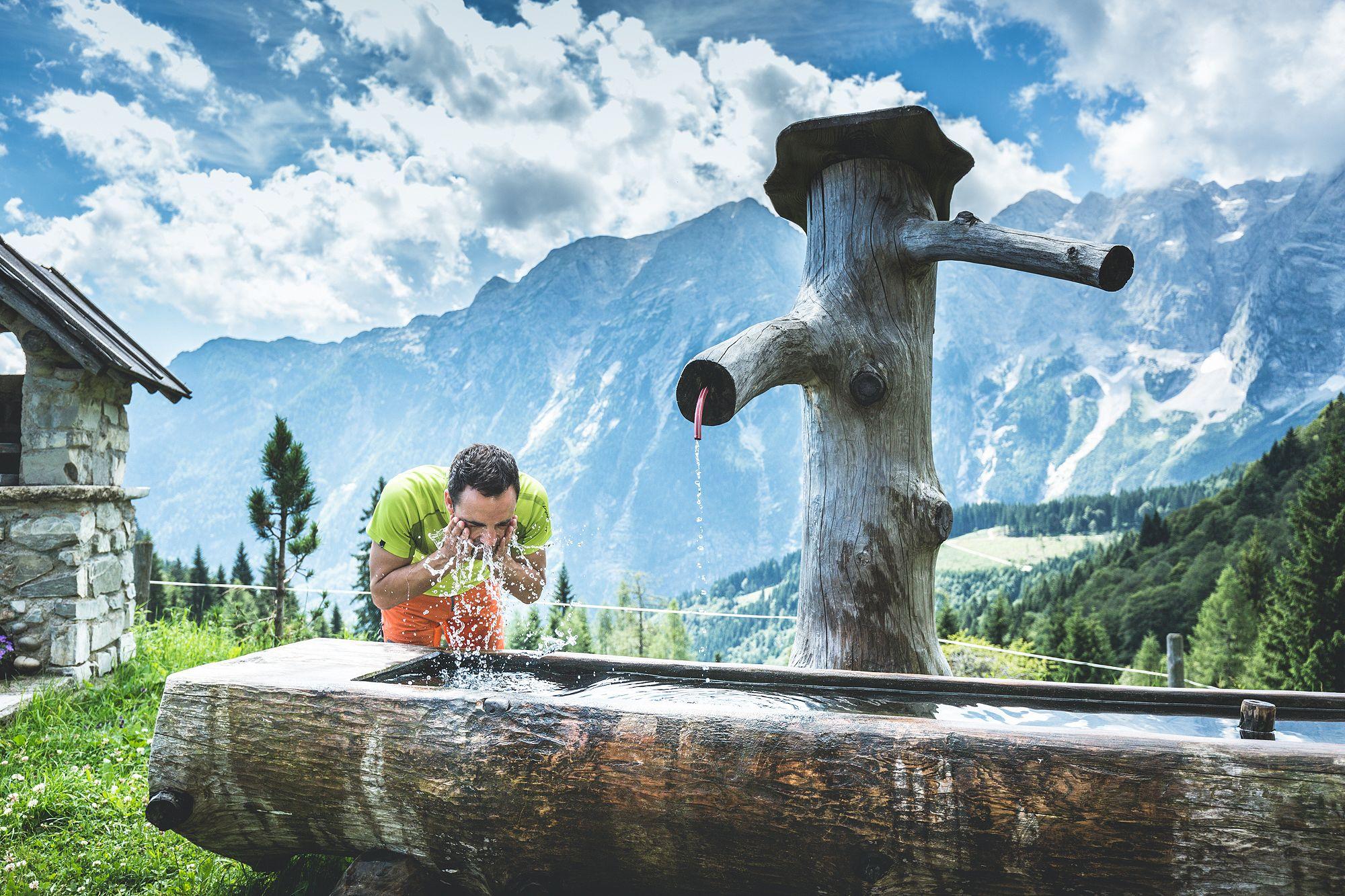 A man drinks fresh water from a wooden well in a picturesque mountain landscape. The surroundings are green and the mountains are visible under a clear sky.
