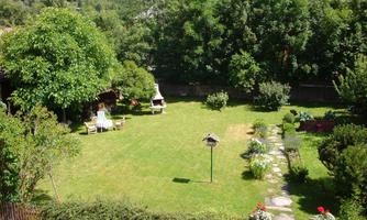 A spacious garden with green grass, lush trees, and colorful flowers. There is a terrace with lounge chairs and a barbecue area.