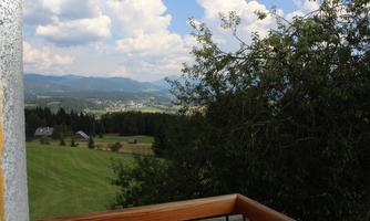 A picturesque view of green fields and mountains under a cloudy sky. In the foreground, a wooden railing can be seen.