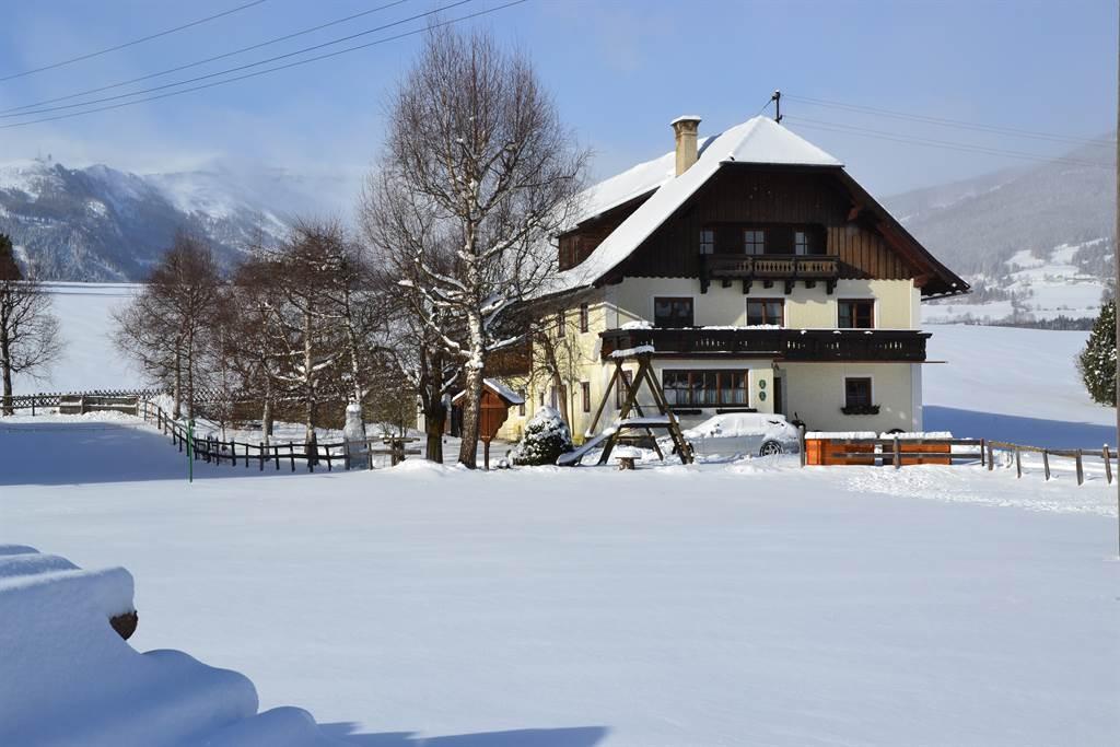 A large, traditional house stands in a snow-covered landscape. The surrounding trees are also covered in snow, and the mountains are visible in the background.