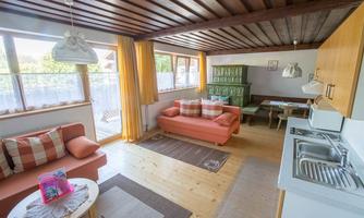 A bright and cozy living and dining room with orange sofas and a dining table. Large windows provide plenty of daylight, and the decor is modern and inviting.