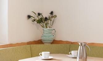 A cozy table in a bright room with a green bench. On the table, there are two cups and a pot, while a vase with flowers decorates the background.