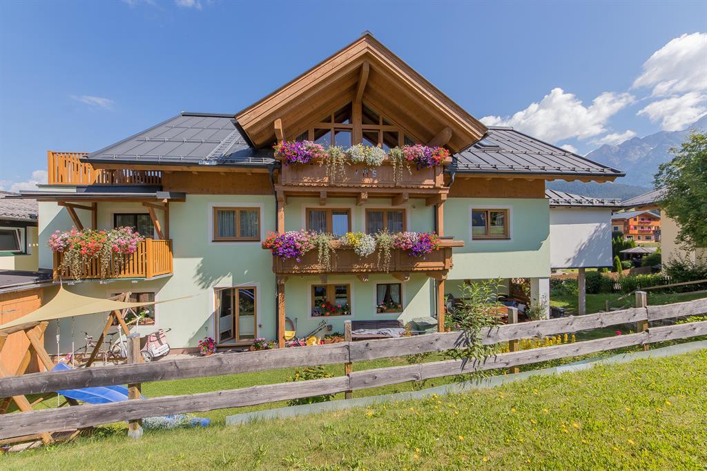 A charming Alpine-style house with blooming balconies. It stands in a green setting under a clear sky.