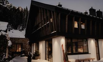 An elegant, modern chalet in a snowy landscape. In the foreground, there are skis, while high mountains rise in the background.
