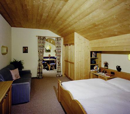 A cozy bedroom with a wooden ceiling and a double bed. In the background, there is a living area with a blue sofa and dining table.