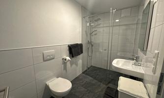 A modern bathroom with a glass shower and elegant white tiles. There is a sink, a toilet, and towels in neutral colors.