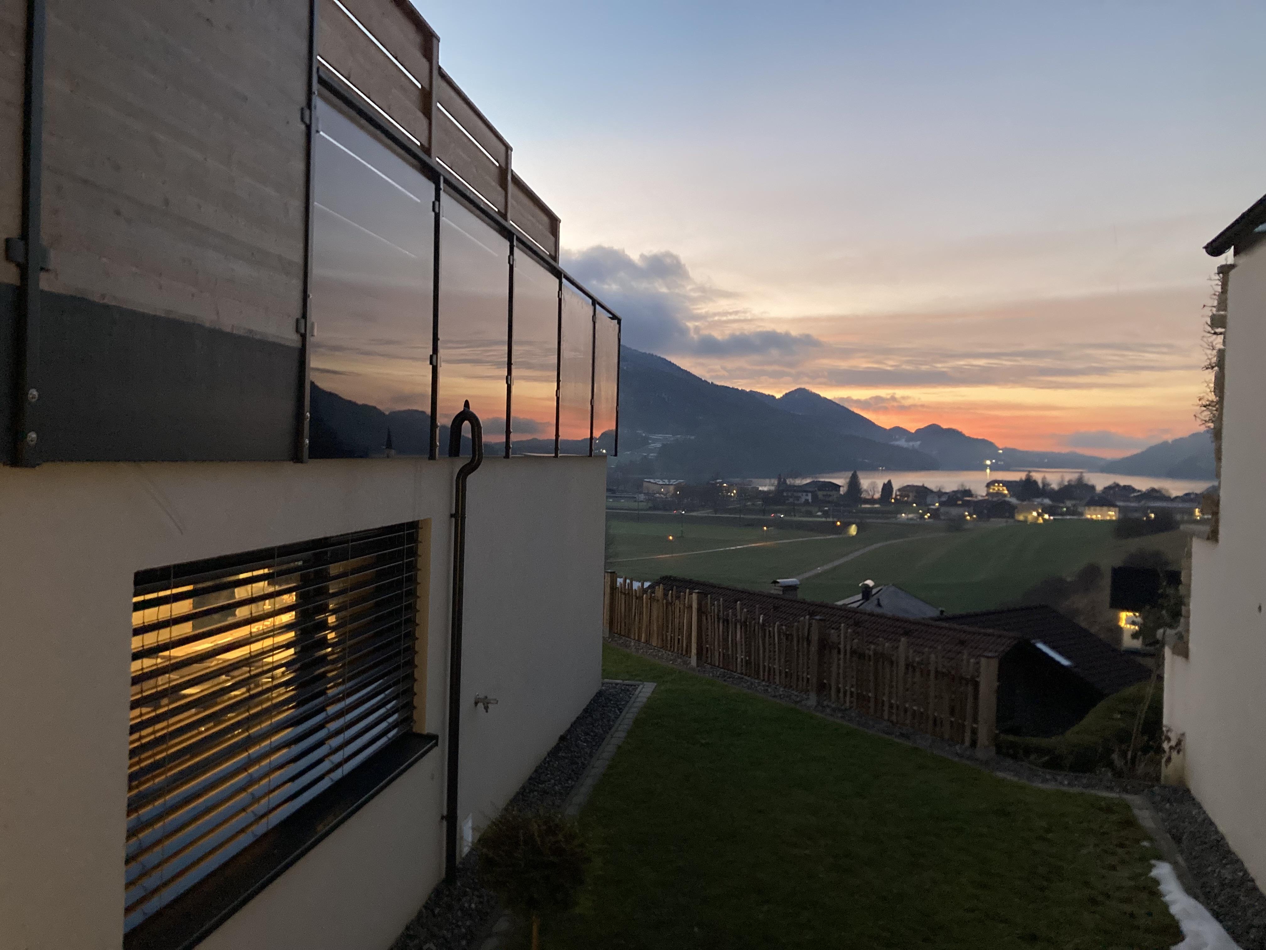 A modern house with large windows overlooks a picturesque landscape. In the background, there are mountains and a gentle sunset.