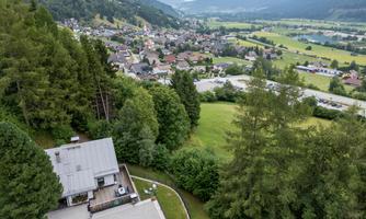 A picturesque landscape with a view of a village in a valley, surrounded by mountains. In the foreground stands a modern house surrounded by trees and meadows.