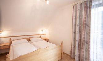 A cozy bedroom with a wooden bed and two mattresses. Large windows with curtains let in plenty of light.