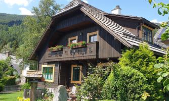 A charming wooden house with flower boxes and a well-maintained garden. The surroundings are green and mountainous, ideal for a quiet retreat.