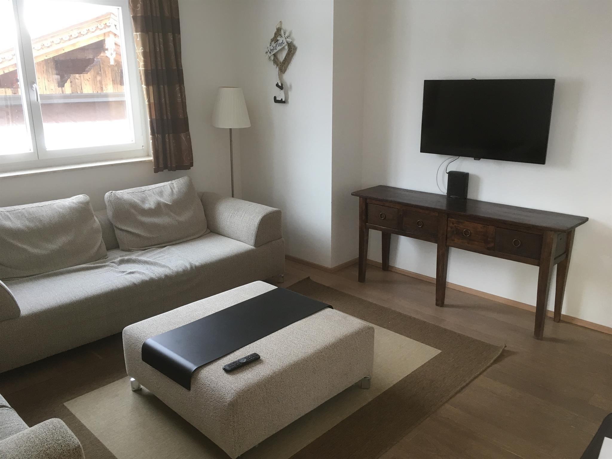 A modern living room with a beige sofa and a coffee table. A television is mounted on the wall, and there is a small wooden table.