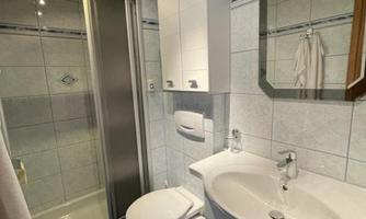 A modern bathroom with a shower, sink, and a toilet. The walls are light tiled and the floor has a dark carpet.