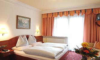 An inviting hotel room with two single beds and a cozy interior. The room has large windows with curtains and a bouquet of flowers on the table.